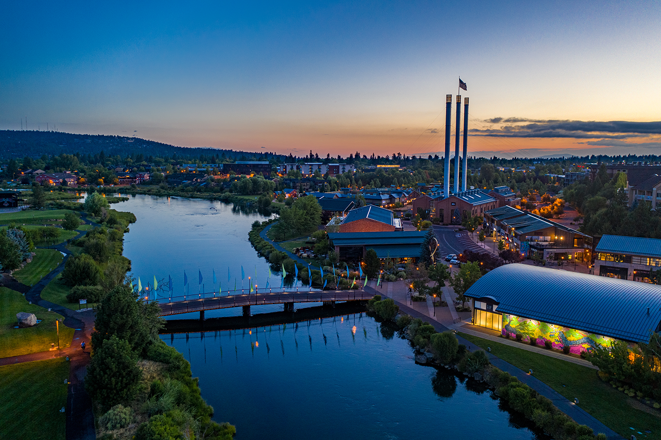 Aerial view of a river running through a town at dusk with a pedestrian bridge, illuminated buildings, and three tall smokestacks with an American flag.