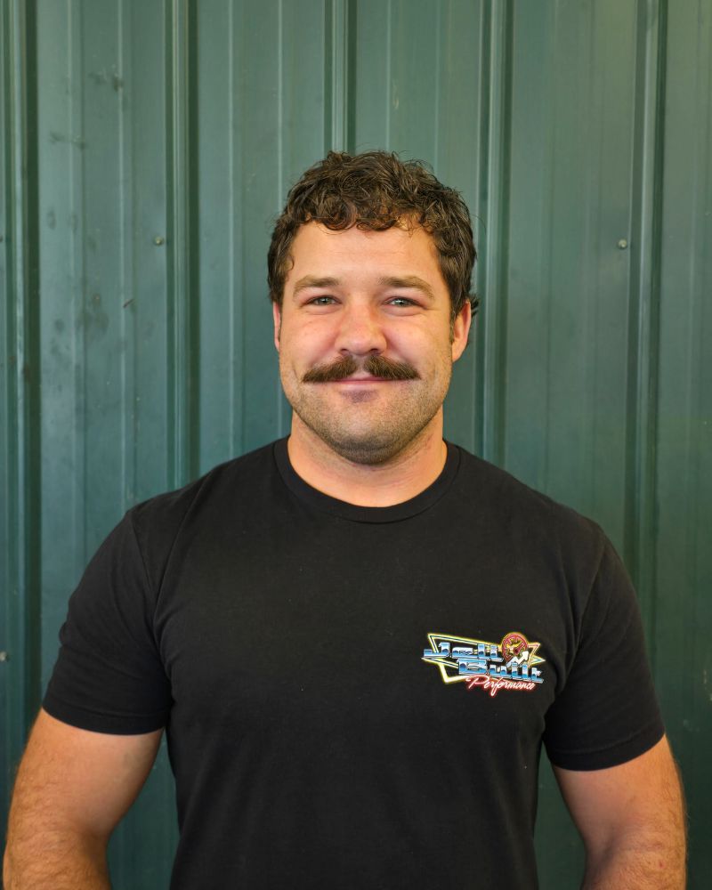 Man with curly hair and mustache wearing a black T-shirt standing against a green metal background.