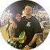 Couple standing close together at an outdoor event with lights in the background, the man holds a yellow rose and a drink.